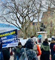 a speaker at a rally stands on a snow-covered lawn before a statue of Benjamin Franklin in front of a University of Pennsylvania building and next to a bare-branched tree; a crowd of people, one person holding a sign saying "HANDS OFF OUR UNIVERSITY" with the logo of the AAUP-Penn chapter,  faces her